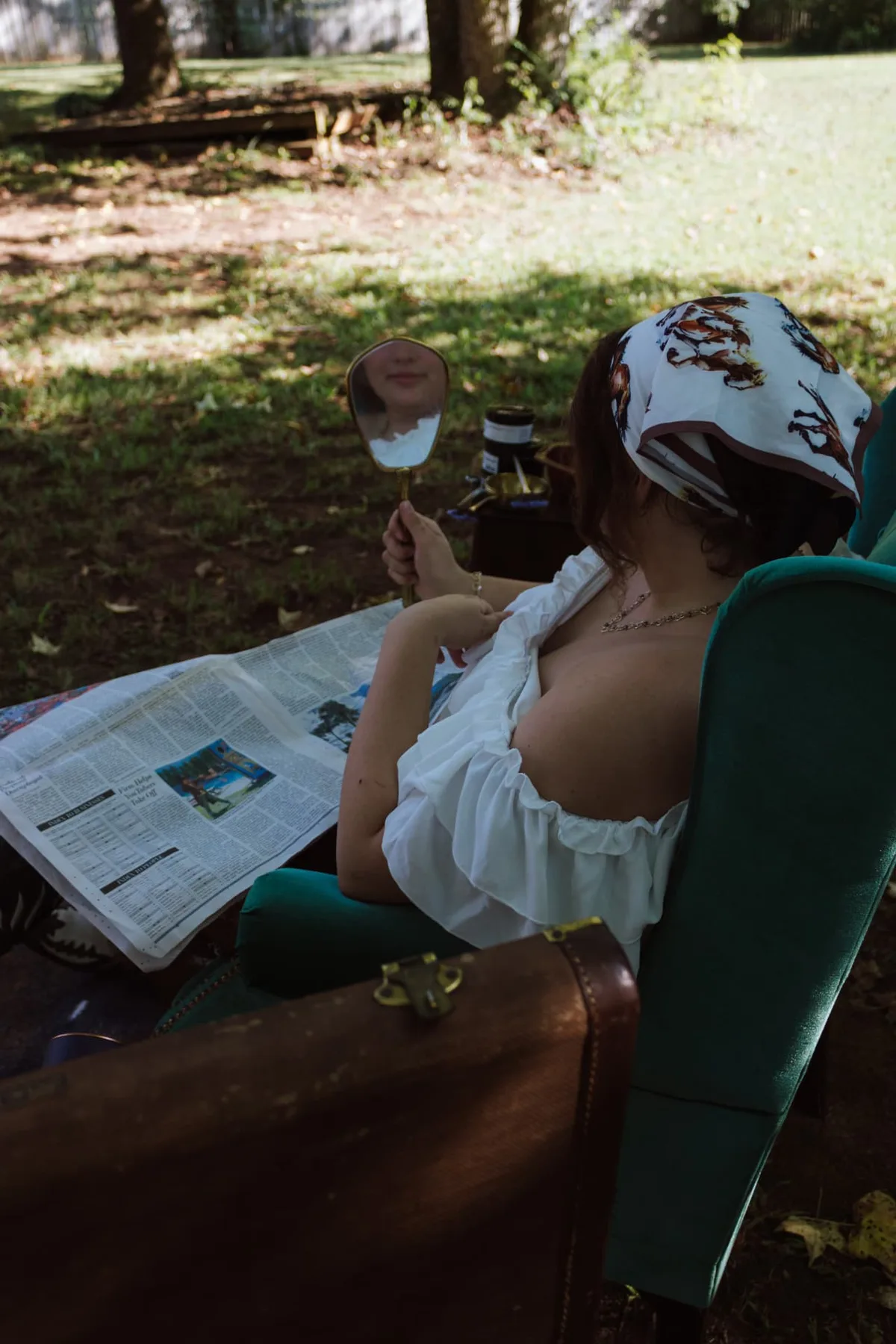 Client relaxing outdoors in a green velvet chair with a hand mirror and newspaper
